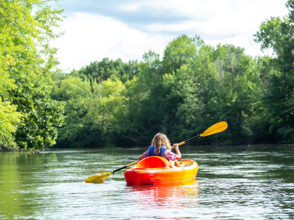 Rear view of a young girl kayaking on the Elk River in Minnesota, USA. She has an orange and black paddle and is sitting in a yellow and orange kayak on this beautiful summer day.